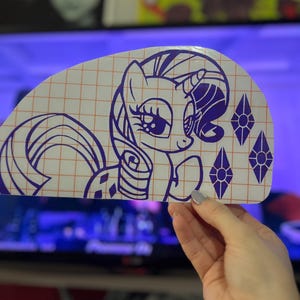 Pu&ograve; includere: Un adesivo in vinile viola di un pony dei cartoni animati con un motivo a rombi sul fianco e un solo rombo sul corno. Il pony guarda a destra e ha un grande occhio espressivo. L'adesivo &egrave; su uno sfondo a griglia.