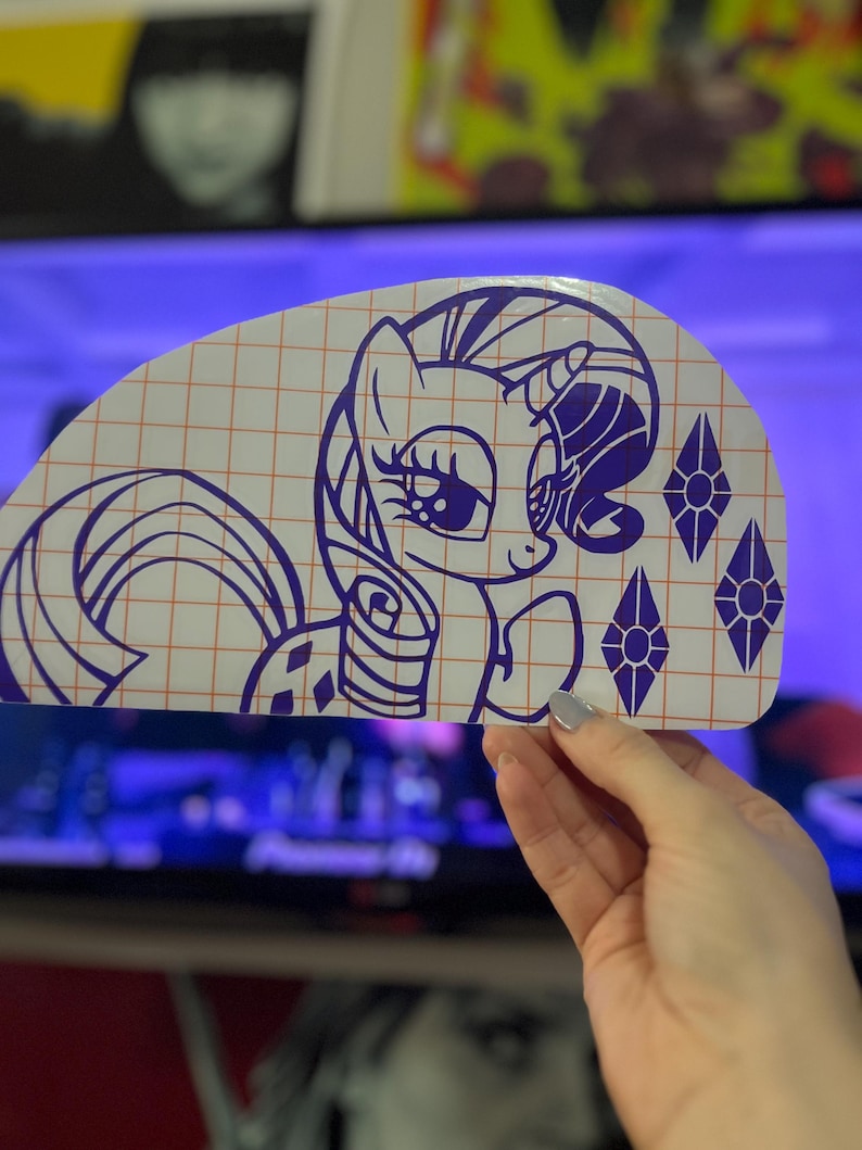 Pu&ograve; includere: Un adesivo in vinile viola di un pony dei cartoni animati con un motivo a rombi sul fianco e un solo rombo sul corno. Il pony guarda a destra e ha un grande occhio espressivo. L'adesivo &egrave; su uno sfondo a griglia.