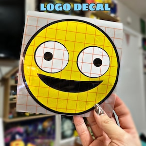 Pode incluir: Um autocolante amarelo com o logótipo Smiling Friends, apresentando um rosto sorridente com olhos pretos e boca preta. O autocolante está sobre um suporte transparente com um padrão de grelha. O texto "SMILING FRIENDS LOGO DECAL" é azul.