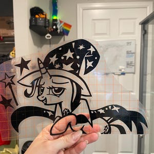 May include: A black vinyl decal featuring a cartoon unicorn wearing a wizard hat decorated with stars. The unicorn holds a star-tipped wand and wears a cape with star patterns. The design is printed on a clear transfer sheet with an orange grid.