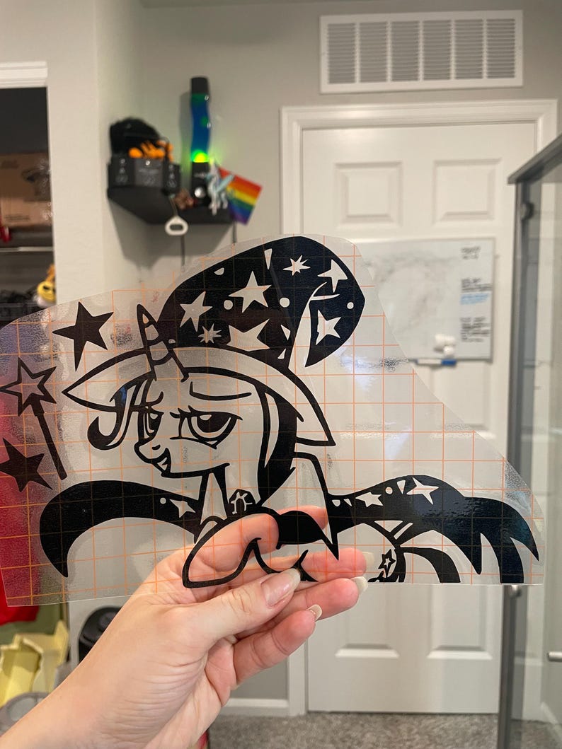 May include: A black vinyl decal featuring a cartoon unicorn wearing a wizard hat decorated with stars. The unicorn holds a star-tipped wand and wears a cape with star patterns. The design is printed on a clear transfer sheet with an orange grid.