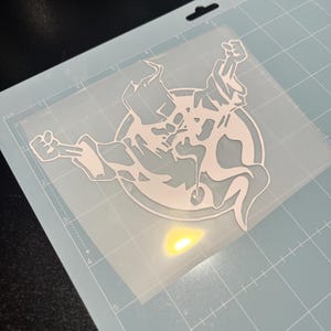 Può includere: Un adesivo in vinile bianco raffigurante una figura spettrale stilizzata con le braccia alzate e una coda fluente. Il disegno è racchiuso in una cornice circolare. L'adesivo è su un tappetino da taglio azzurro.