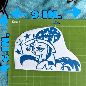 May include: A blue vinyl decal featuring a cartoon unicorn wearing a wizard hat decorated with stars, holding a magic wand. The decal is positioned on a green cutting mat, with text indicating dimensions of 9 inches by 6 inches for the design.