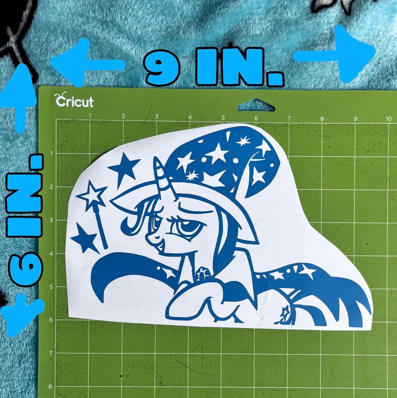 May include: A blue vinyl decal featuring a cartoon unicorn wearing a wizard hat decorated with stars, holding a magic wand. The decal is positioned on a green cutting mat, with text indicating dimensions of 9 inches by 6 inches for the design.