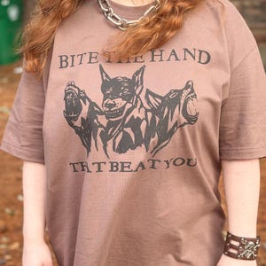 May include: A brown t-shirt featuring the text "BITE THE HAND THAT BEAT YOU" and a graphic of three snarling dogs. The shirt has short sleeves and is worn with a silver chain necklace and a brown leather bracelet with skull accents.