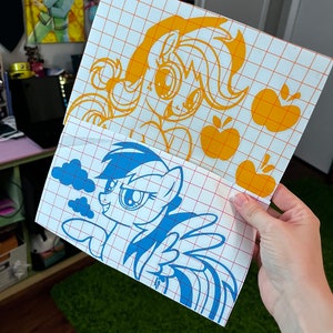 May include: Two vinyl decals featuring cartoon ponies. The top decal is yellow and features a pony with a yellow mane and tail. The bottom decal is blue and features a pony with a blue mane and tail. Both decals have a grid pattern behind them.