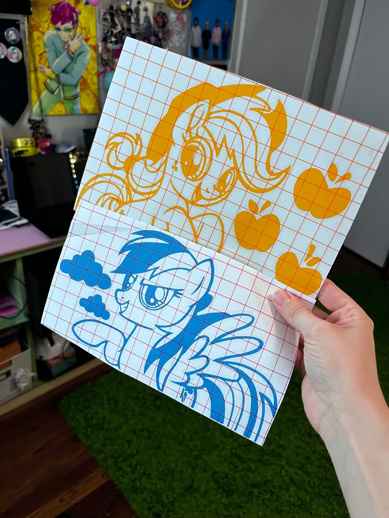May include: Two vinyl decals featuring cartoon ponies. The top decal is yellow and features a pony with a yellow mane and tail. The bottom decal is blue and features a pony with a blue mane and tail. Both decals have a grid pattern behind them.
