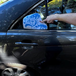 May include: A blue vinyl decal featuring a cartoon unicorn wearing a wizard hat with stars, applied to the rear passenger window of a dark gray car. The unicorn has a horn, a flowing mane, and a star-patterned tail, with small stars nearby.