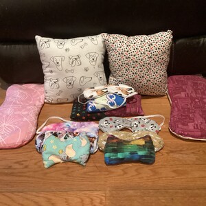 May include: A collection of eight pillows and eye masks in various colors and patterns. The pillows are rectangular and feature different designs, including dogs, flowers, and geometric shapes. The eye masks are made of soft fabric and have different colors and patterns.