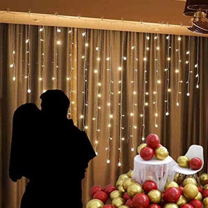 May include: A couple silhouetted in front of a curtain of white fairy lights. The lights are strung in a cascading pattern. A table with a white tablecloth is decorated with red and gold balloons.