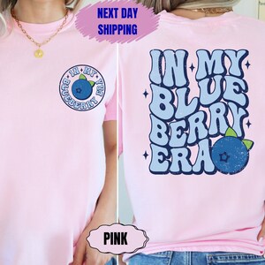 May include: Pink t-shirt with a graphic on the back that says "In My Blue Berry Era" in a retro font with a blueberry graphic. The front of the shirt has a smaller graphic of a blueberry in a circle with the text "In My Blueberry Era".