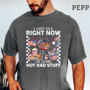May include: A gray t-shirt with a graphic that reads "I CAN'T TALK RIGHT NOW I'M DOING HOT DAD STUFF." The graphic features a barbecue grill, American flags, and food items like hot dogs and burgers. The shirt is a casual style.