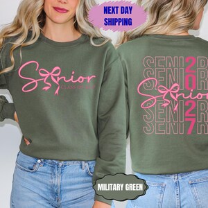 May include: Military green sweatshirt with the word "Senior" in pink script on the front, with a bow design. The back of the sweatshirt repeats "Senior 2027" in a stacked design. The sweatshirt is a crew neck style.