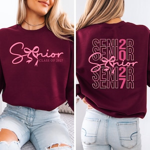 May include: Burgundy sweatshirt with the word "Senior" in pink script and a bow design. The text "Class of 2027" is below the word "Senior". The back of the sweatshirt has the word "Senior" repeated with the numbers "2027".