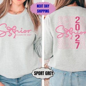 May include: A light gray sweatshirt with the word "Senior" in pink script with a bow design. The text "Class of 2027" is below. The back of the sweatshirt has the word "Senior" and the number "2027" repeated in a vertical pattern.