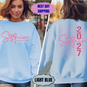 May include: Light blue sweatshirt with the word "Senior" in pink script with a bow design. The back of the sweatshirt also features the word "Senior" and the number "2027" in pink. The sweatshirt is a light blue color.