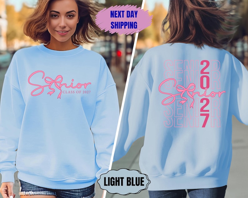 May include: Light blue sweatshirt with the word "Senior" in pink script with a bow design. The back of the sweatshirt also features the word "Senior" and the number "2027" in pink. The sweatshirt is a light blue color.