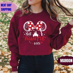 May include: Maroon sweatshirt with a white graphic of a bow, mouse ears, and the text "Best Auntie Ever." The design includes a castle and hearts. The sweatshirt also has a "Next Day Shipping" and "Maroon" label.