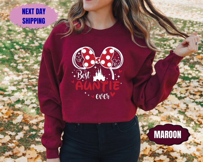May include: Maroon sweatshirt with a white graphic of a bow, mouse ears, and the text "Best Auntie Ever." The design includes a castle and hearts. The sweatshirt also has a "Next Day Shipping" and "Maroon" label.