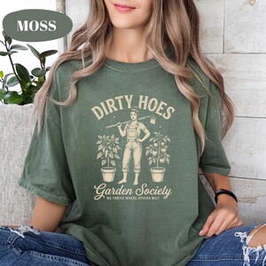 May include: A moss green t-shirt with the words "DIRTY HOES" above a graphic of a person holding a hoe, flanked by potted plants. Below, the text reads "Garden Society" and "WE THRIVE WHERE OTHERS WILT".