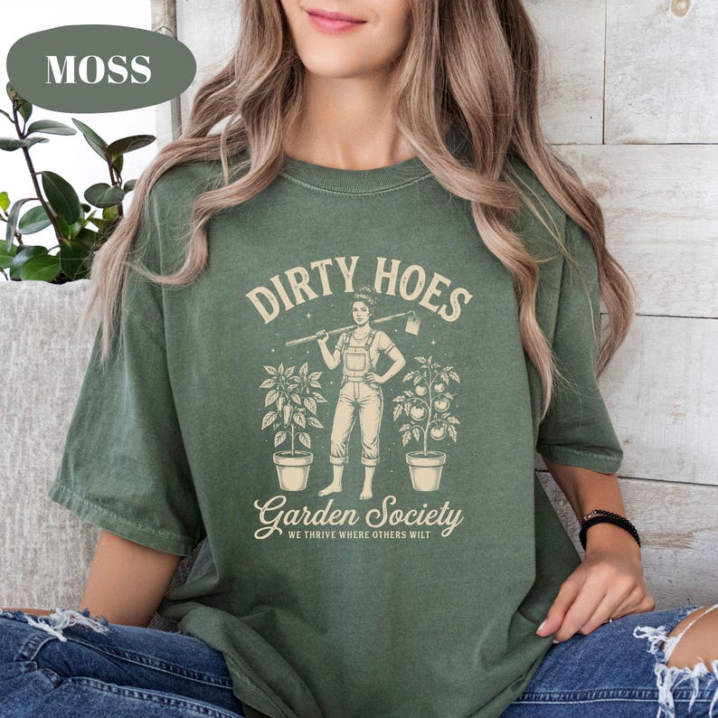 May include: A moss green t-shirt with the words "DIRTY HOES" above a graphic of a person holding a hoe, flanked by potted plants. Below, the text reads "Garden Society" and "WE THRIVE WHERE OTHERS WILT".