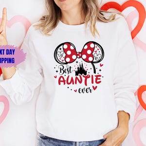 May include: White sweatshirt with a graphic that says "Best Auntie ever." The graphic includes a red polka dot bow, a black castle, and red hearts. The sweatshirt is long-sleeved.
