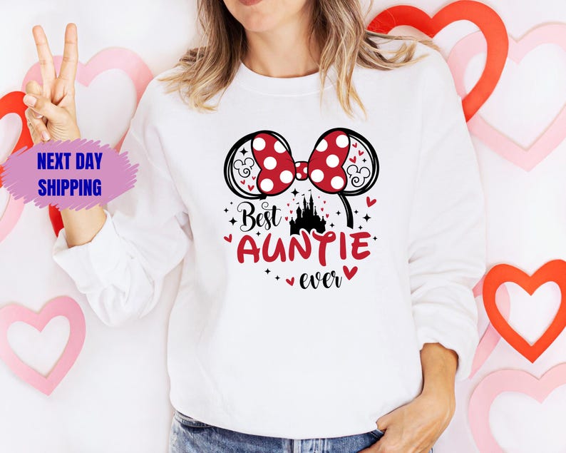 May include: White sweatshirt with a graphic that says "Best Auntie ever." The graphic includes a red polka dot bow, a black castle, and red hearts. The sweatshirt is long-sleeved.