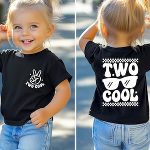 May include: Black toddler t-shirt with white graphics. The front features a peace sign and the words "TWO COOL". The back displays "TWO COOL" with sunglasses and a checkered pattern.