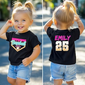 May include: A black t-shirt with a baseball-themed design on the front, featuring the words "Baseball Sister" and a baseball graphic. The back of the shirt displays the name "Emily" and the number "25". The shirt is paired with denim shorts.