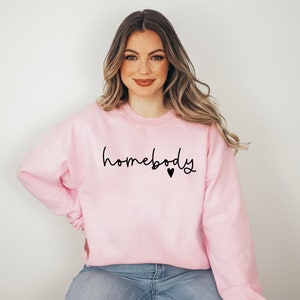 May include: A pink sweatshirt with the text "homebody" in black script with a small black heart.