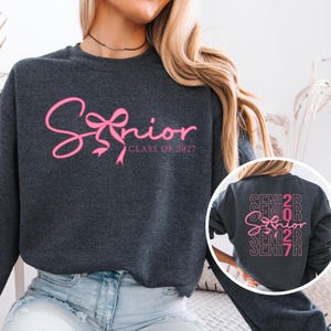 May include: A dark gray sweatshirt with the word "Senior" in pink script, accented with a bow design. The text "Class of 2027" is below. The back of the sweatshirt has the word "Senior" repeated.