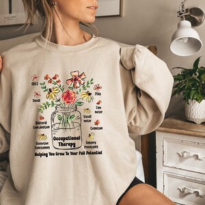 May include: A beige sweatshirt with a floral design and the text "Occupational Therapy" in a jar. The flowers represent different aspects of occupational therapy, such as ADLs, social skills, fine motor skills, visual motor skills, cognition, executive functioning, and sensory processing. The text below the jar reads "Helping You Grow To Your Full Potential."