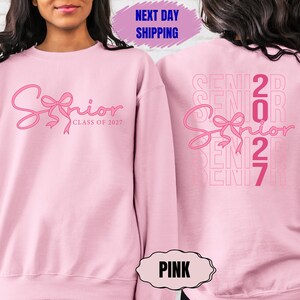 May include: Pink sweatshirt with the word "Senior" in a cursive font and a bow design. The text "Class of 2027" is below the word "Senior". The back of the sweatshirt has the word "Senior" and the numbers "2027".