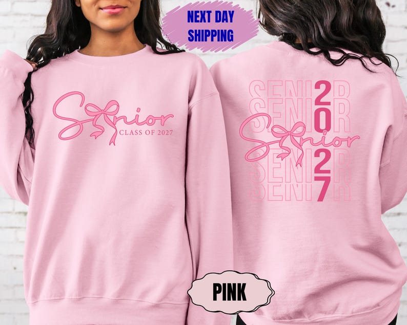 May include: Pink sweatshirt with the word "Senior" in a cursive font and a bow design. The text "Class of 2027" is below the word "Senior". The back of the sweatshirt has the word "Senior" and the numbers "2027".