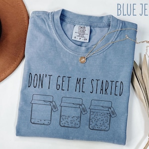 May include: Blue jean Comfort Colors t-shirt with the phrase "DON'T GET ME STARTED" and three jar illustrations. A gold necklace with a pendant is draped across the shirt. The shirt is folded on a white surface.