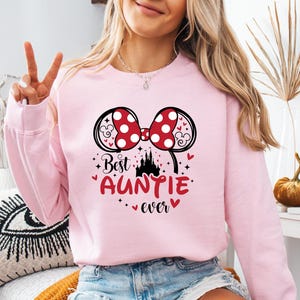 May include: A light pink sweatshirt with a graphic that says "Best Auntie Ever." The graphic includes a red bow with white polka dots, black mouse ears, a castle, and hearts.