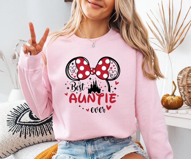 May include: A light pink sweatshirt with a graphic that says "Best Auntie Ever." The graphic includes a red bow with white polka dots, black mouse ears, a castle, and hearts.