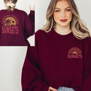 May include: A maroon sweatshirt with a graphic on the front and back. The front graphic is a stylized sunset with palm trees and the word "SUNSETS". The back graphic is a circle with the words "FOREVER CHASING SUNSETS" and a palm tree.