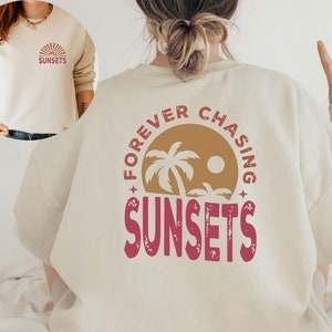May include: A cream colored sweatshirt with the text "FOREVER CHASING SUNSETS" in a circle with a palm tree graphic. The text is in a red and brown color scheme.