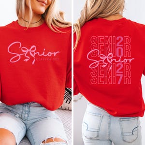 May include: Red sweatshirt with the word "Senior" in pink script and a bow graphic. The front of the sweatshirt also includes "Class of 2027". The back of the sweatshirt repeats "Senior" in a stacked design with the year.