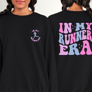 May include: Black sweatshirt with a pink and blue graphic on the back that says "IN MY RUNNER ERA" and a smaller pink and blue graphic on the front that says "IN MY RUNNER ERA" with a running figure.