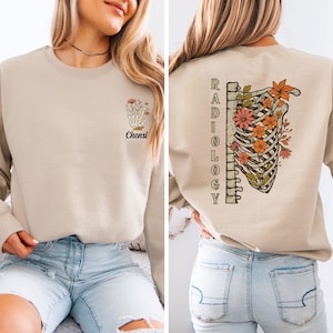 May include: Beige sweatshirt with a floral skeleton hand design on the front and the name "Chensi" below it. The back features a rib cage design filled with flowers and the word "Radiology" vertically.
