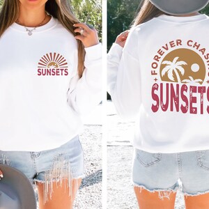 May include: White long-sleeve sweatshirt with a red graphic on the front that says "SUNSETS" with a stylized sun and palm trees. The back of the sweatshirt has a red graphic that says "FOREVER CHASING SUNSETS" with a stylized sun and palm trees.