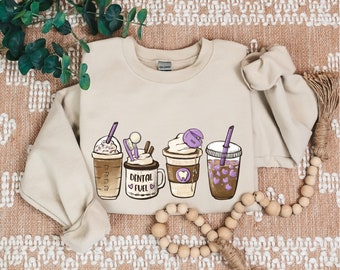 May include: A beige sweatshirt featuring a graphic of four coffee cups with purple accents. The cups are labelled "Dental Fuel".