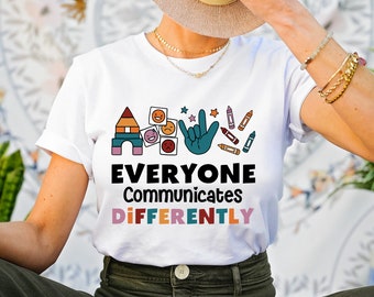 May include: White t-shirt with a colorful graphic of building blocks, crayons, and a hand signing "I love you". The text "Everyone Communicates Differently" is printed below the graphic.