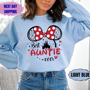 May include: Light blue sweatshirt featuring a Minnie Mouse bow with red polka dots and the text "Best Auntie ever" in red. The design includes a castle and hearts. The sweatshirt is a casual style.