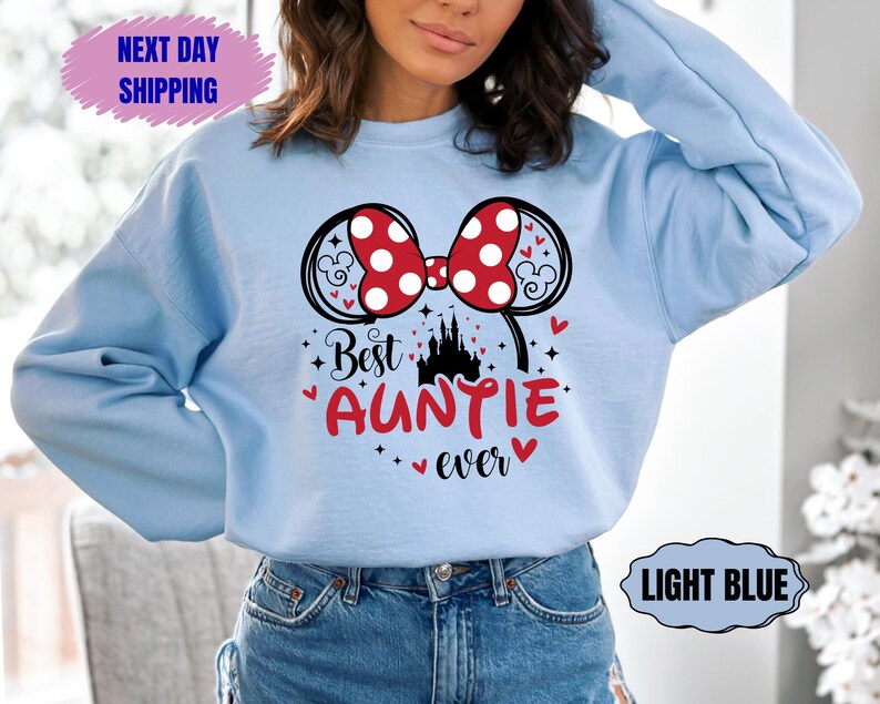 May include: Light blue sweatshirt featuring a Minnie Mouse bow with red polka dots and the text "Best Auntie ever" in red. The design includes a castle and hearts. The sweatshirt is a casual style.