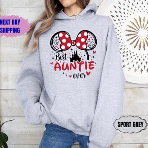 May include: Sport grey hoodie with a Minnie Mouse bow design in red and white polka dots. The text "Best Auntie ever" is in red with a castle graphic. The hoodie has a drawstring hood and a front pocket.