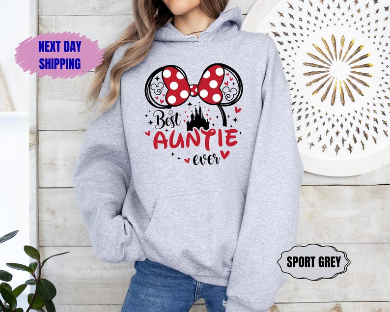 May include: Sport grey hoodie with a Minnie Mouse bow design in red and white polka dots. The text "Best Auntie ever" is in red with a castle graphic. The hoodie has a drawstring hood and a front pocket.