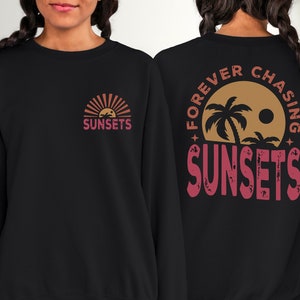May include: Black sweatshirt with a graphic of a sunset and palm trees on the back. The text "FOREVER CHASING SUNSETS" is printed in red on the back of the sweatshirt. The front of the sweatshirt has a smaller graphic of a sunset with the word "SUNSETS" printed in red.